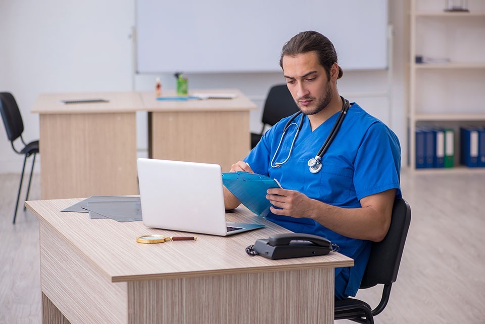 medical scribe wearing a stethoscope sits at a computer and reviews patient notes, typing and referencing documents in a clinic setting