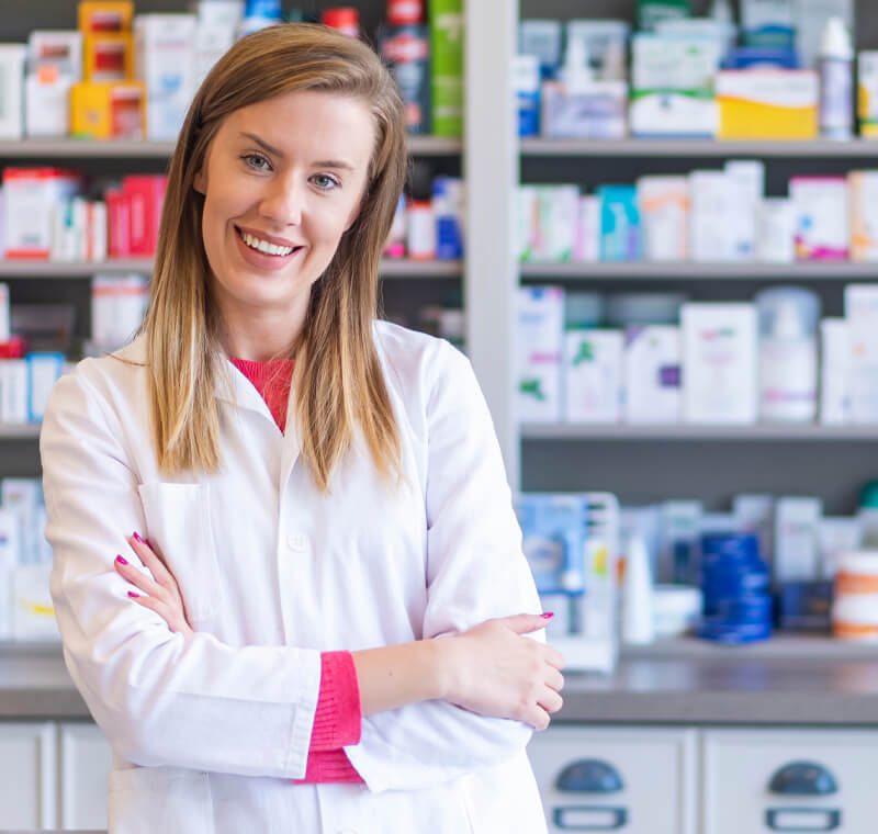 A pharmacy technician specialist in a white lab coat stands confidently in a well-stocked pharmacy, organizing medication bottles and consulting a computer screen.